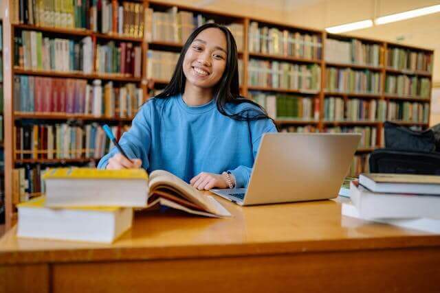 A student in a library studying the differences between German dative and accusative cases with books and a laptop