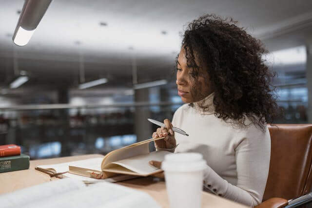 A woman studying in a library, writing notes to understand the difference between 'ob' and 'wenn' in German grammar.