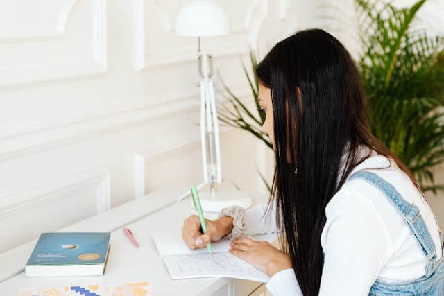 A young woman studying German Dative Case rules at a desk, writing in a notebook with focus.