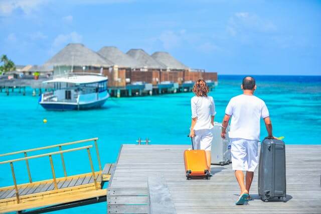 Couple walking on a dock with luggage towards overwater villas, symbolizing German Travel Vocabulary.