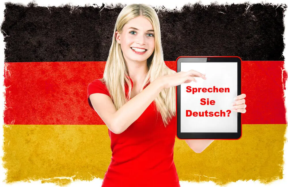 German teacher teaching German language holding a tablet with the text 'Sprechen Sie Deutsch?' in front of a German flag background.