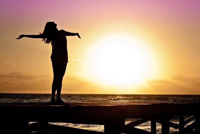 Silhouette of a woman with open arms at sunset symbolizing joy and freedom using German nouns vocabulary for emotions and feelings.