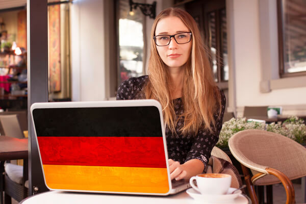 Young woman working on a laptop with a German flag design, practicing the perfect tense in German at an outdoor café.