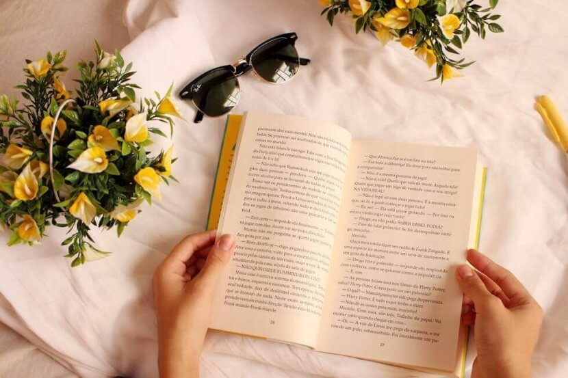 Hands holding an open book surrounded by flowers and sunglasses, symbolizing the joy of learning German literature and books vocabulary