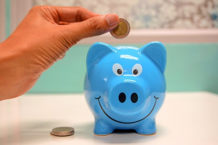 Hand placing a coin into a blue piggy bank, representing German banking vocabulary like Geld sparen (save money).