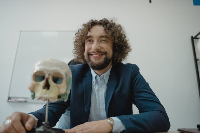 Man with curly hair smiling at a desk with a human skull model, representing German Body Parts Vocabulary.