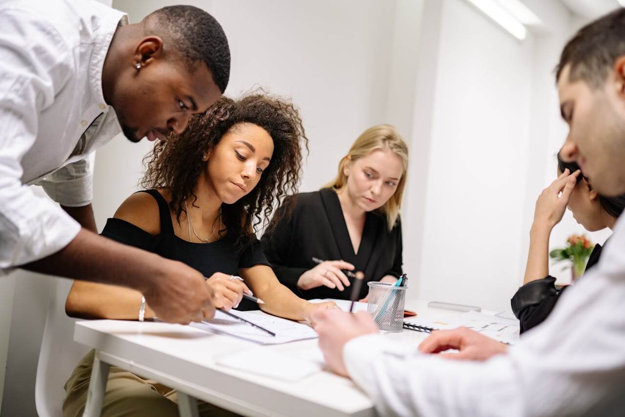A diverse team of colleagues gathers around a table to discuss and plan das Projekt (the project), focusing intently on their documents while collaborating in a bright office space.
