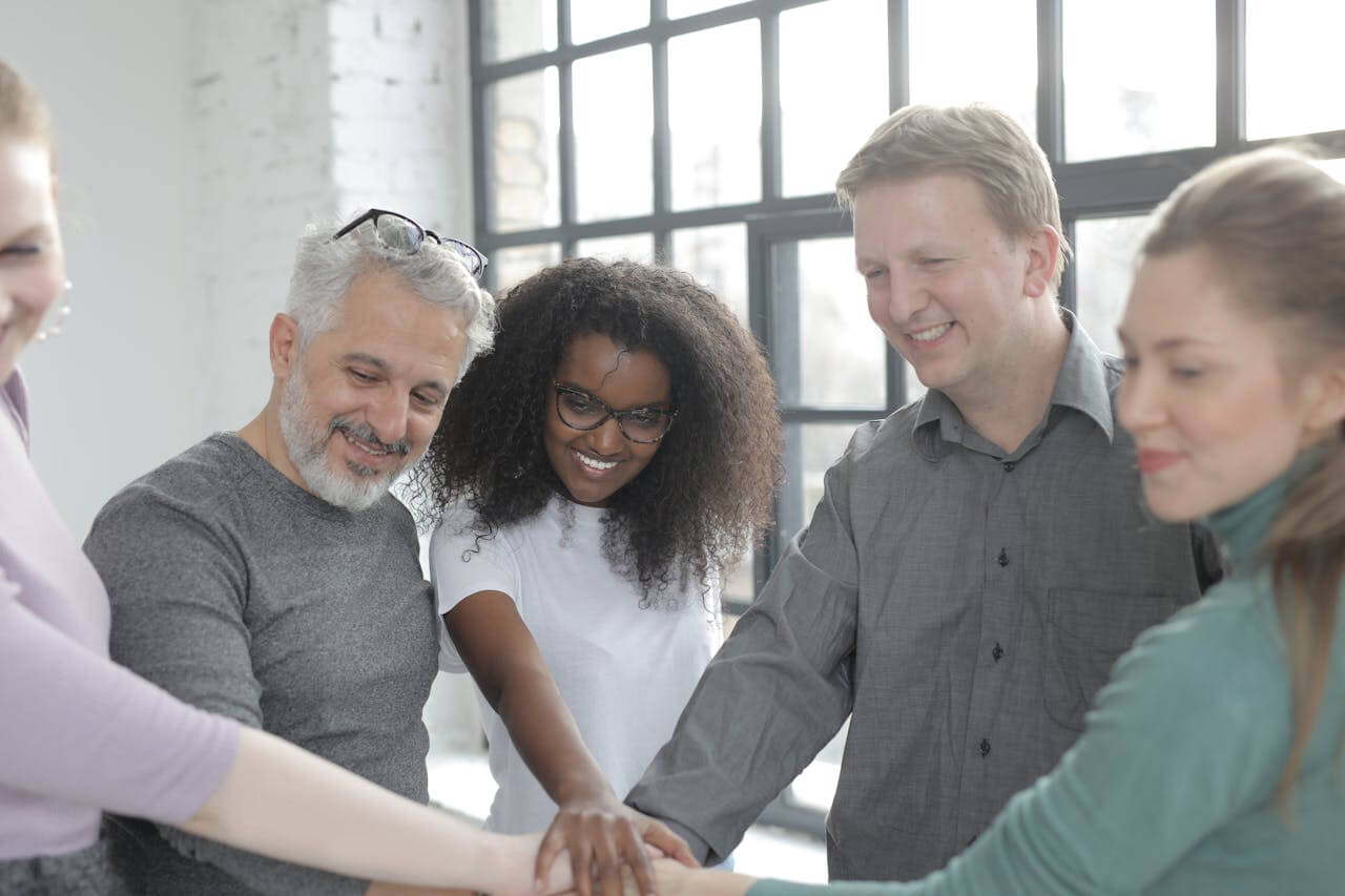 A group of smiling colleagues, of diverse ages and backgrounds, place their hands together in a central stack, signifying unity and joint effort, showing their commitment to das Team (the team) as they work towards a common goal in a modern office.