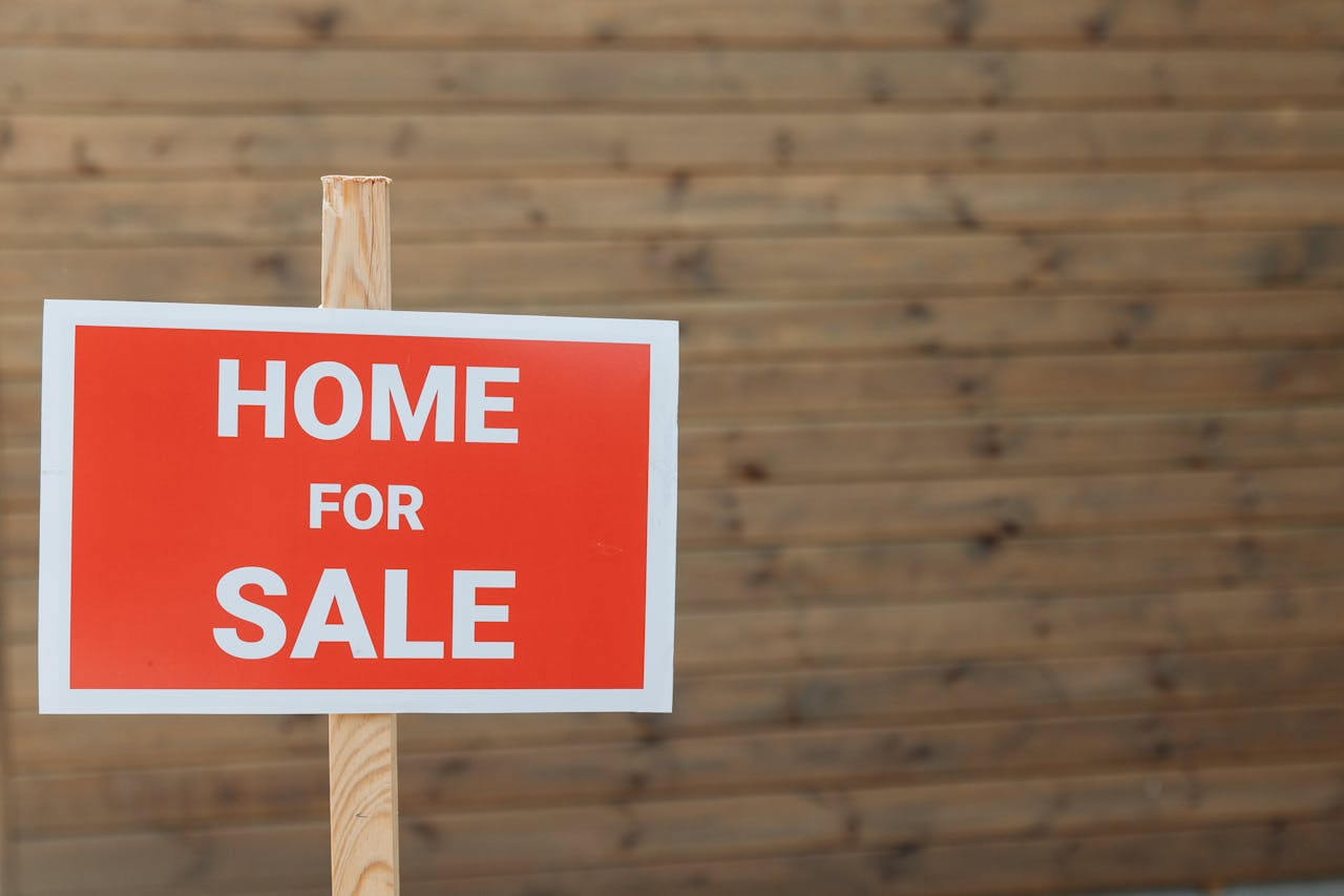 A bright red 'HOME FOR SALE' sign is mounted on a wooden post against a rustic wooden background, signaling the start of der Verkauf (the sale) for a piece of real estate.