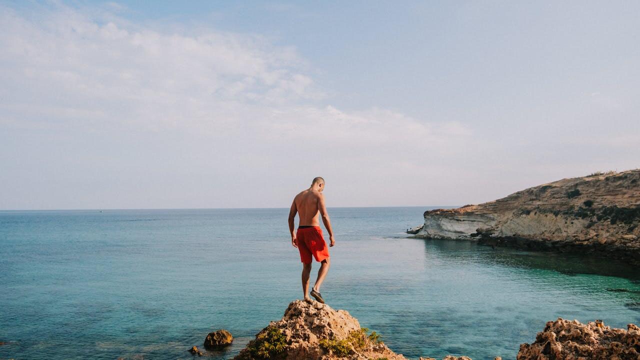 A man stands on a rocky cliff overlooking a beautiful turquoise sea and wears die Badehose (the swimming trunks) in a bright red color, looking down at the clear water while enjoying a sunny day at the coast.