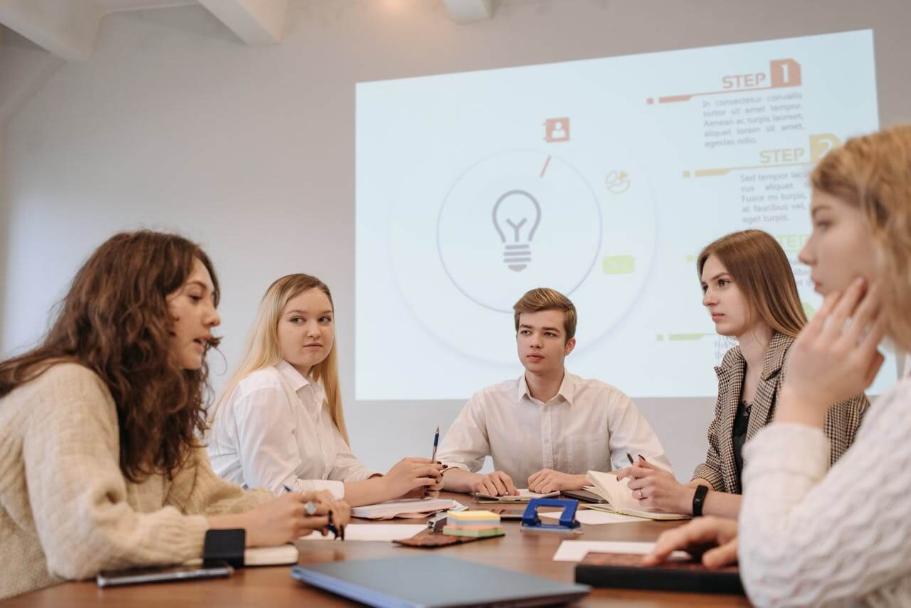 A group of colleagues sit together in die Besprechung (the meeting), discussing ideas while looking at notes and a presentation on the screen.