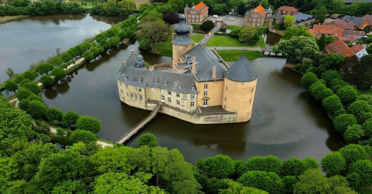 Aerial view of the historic moated castle Burg Vischering in Germany surrounded by a moat and lush green trees, serving as the hero image for the grammar guide on the German noun 'der Neffe'.