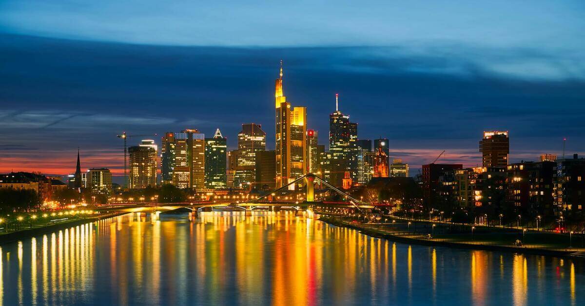 Illuminated skyscrapers of the Frankfurt skyline reflected in the river Main at night, serving as the hero image for the grammar guide on the German noun 'die Mutter'.