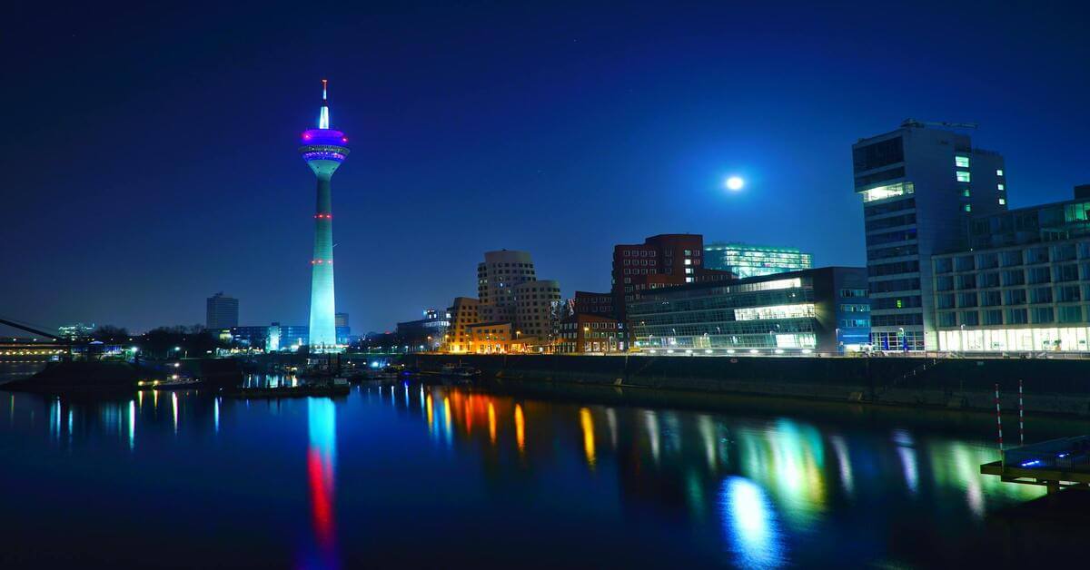Night view of the illuminated Dusseldorf Medienhafen featuring the Rhine Tower and other modern buildings, serving as the hero image for the grammar guide on the German noun 'die Schwester'.