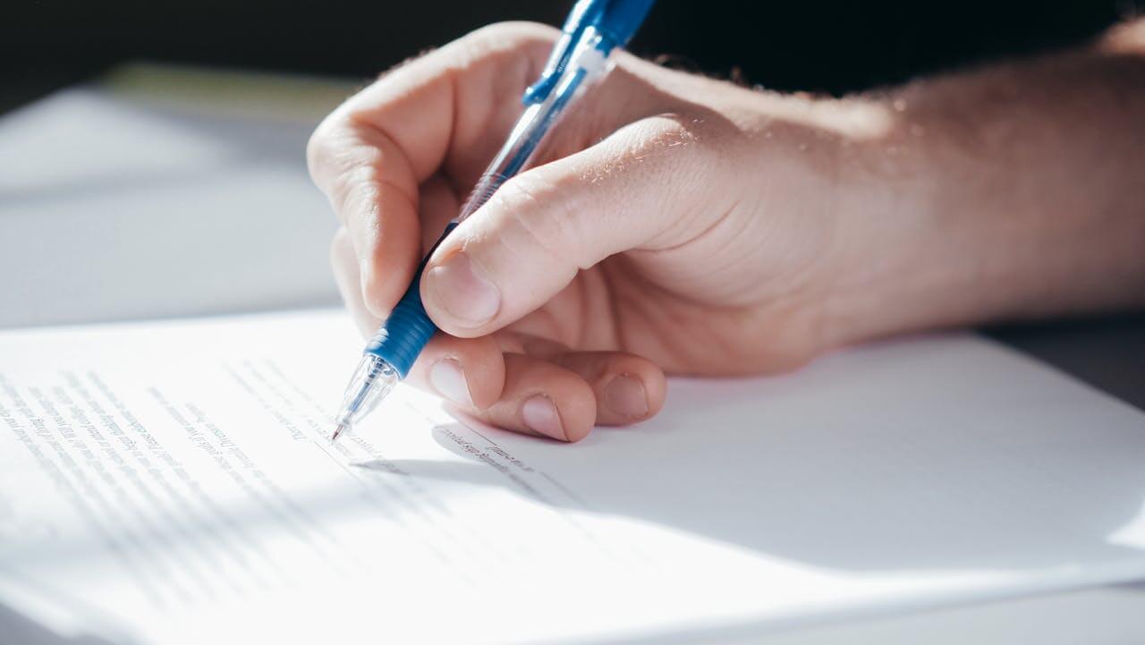 Close-up shot of a person's hand signing a document with a blue pen, illustrating the German masculine noun "der Arbeitsvertrag" (the employment contract).