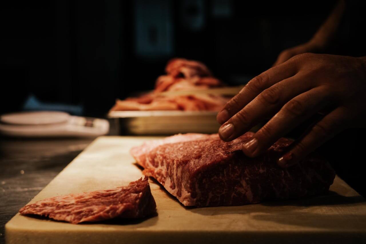 A chef’s hand rests on a high-quality, marbled piece of raw beef on a cutting board in a professional kitchen, preparing das Fleisch (the meat) which serves as a nutrient-dense main ingredient for many traditional and modern dishes.