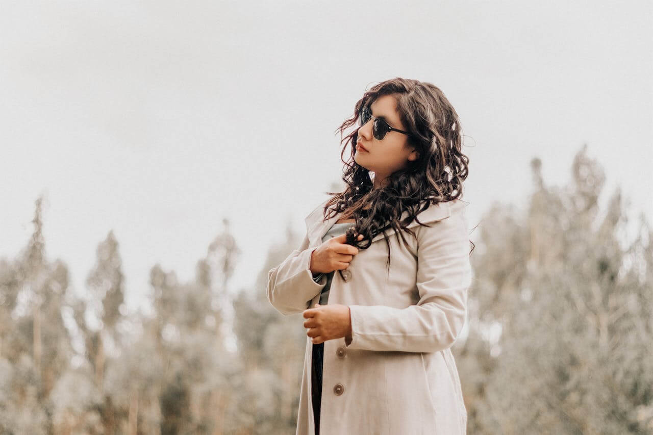 A woman with long curly hair and sunglasses stands outdoors against a soft-focus forest background, wearing a light-colored der Mantel (the coat), which serves as a stylish outer layer to protect against the wind and cold.
