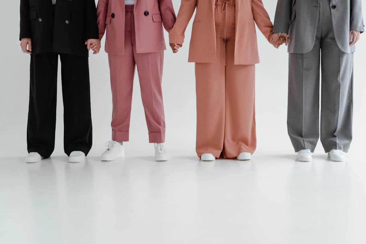 Four people stand in a row holding hands, each wearing a different style and color of die Hose (the pants), which serves as a versatile garment to cover the legs and complete a stylish outfit.