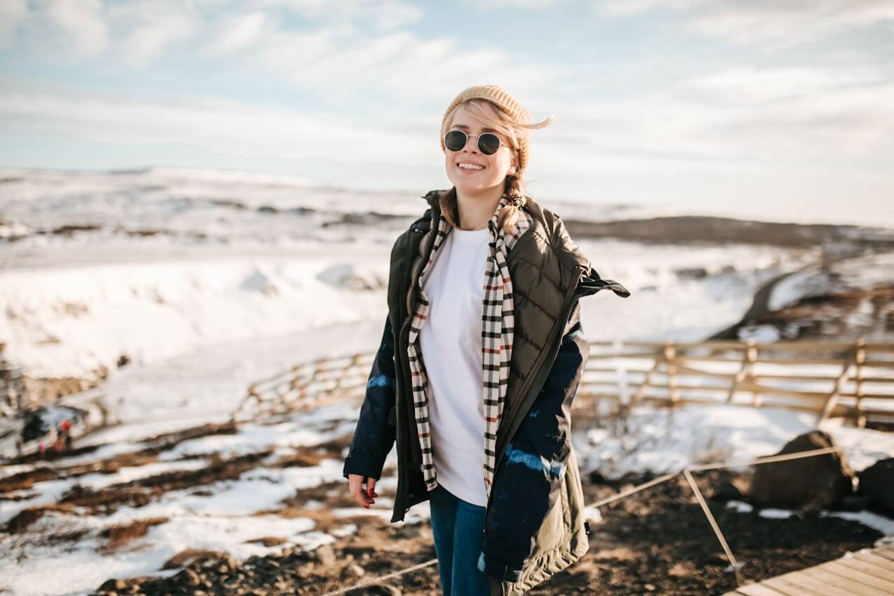 A smiling young woman, wearing a beanie and sunglasses, walks on a wooden boardwalk through a snowy, rocky landscape, wrapped in a layered olive-green and blue die Jacke (the jacket) to keep her warm against the elements.
